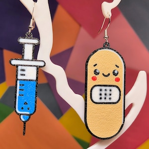 May include: A pair of earrings with a blue syringe and a cartoon band-aid. The band-aid has a smiling face with red cheeks and a white bandage.