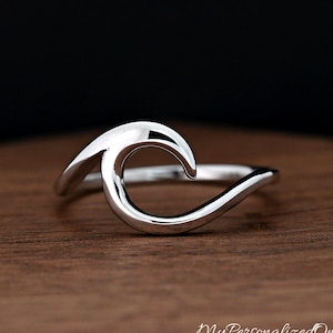 May include: A silver-colored ring in the shape of a wave. The ring is smooth and polished, with a curved design that mimics the crest of a wave. The ring is set against a wooden surface.