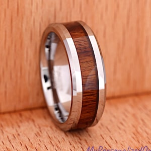 May include: A polished silver-toned metal ring featuring a wide, flat inlay of dark brown wood grain running horizontally around the center of the band. The metal edges are beveled and reflective, creating a contrast with the natural wood texture.