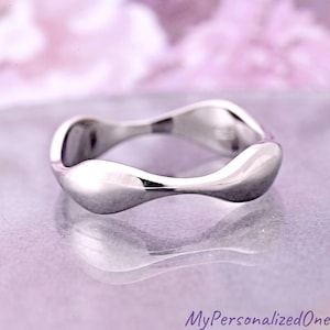 May include: A polished silver ring with a flowing, wave-like design. The ring has a smooth, reflective surface and a modern, minimalist aesthetic. The ring is displayed on a reflective surface with a soft, blurred background.