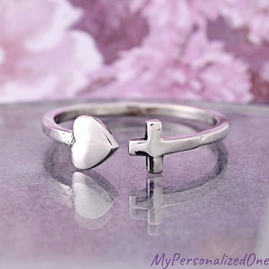 May include: A silver-toned open ring featuring a polished heart shape on one end and a small cross symbol on the other end. The band is smooth and metallic, designed with an open gap between the two distinct charms, creating a simple and reflective piece of jewelry.