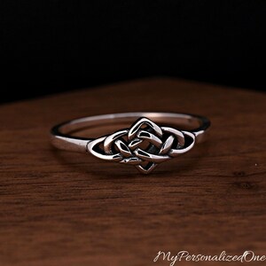 May include: A silver ring featuring a central knot design with polished surfaces and darkened recessed areas. The band is smooth and circular, showcasing a continuous metal loop that forms an intricate, symmetrical pattern at the front centre.