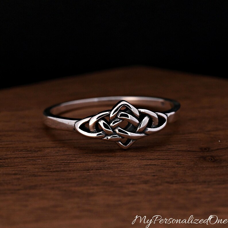 May include: A silver ring featuring a central knot design with polished surfaces and darkened recessed areas. The band is smooth and circular, showcasing a continuous metal loop that forms an intricate, symmetrical pattern at the front centre.