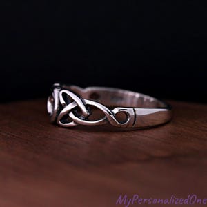 May include: A polished silver ring featuring an intricate, continuous triquetra knot design. The band has a smooth, rounded profile with darkened recessed areas that highlight the woven metal pattern, creating a clear contrast against the bright silver.