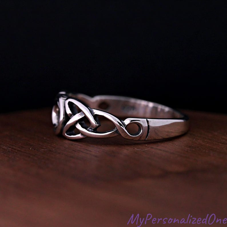 May include: A polished silver ring featuring an intricate, continuous triquetra knot design. The band has a smooth, rounded profile with darkened recessed areas that highlight the woven metal pattern, creating a clear contrast against the bright silver.