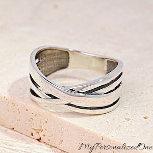 May include: A silver-toned ring featuring a multi-band design with overlapping, crisscrossing segments that create a layered appearance. The metal surface has a polished finish with darkened recessed areas between the bands to add depth.