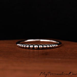May include: A silver ring with a beaded design. The ring features a row of small, evenly spaced beads along the top, creating a textured appearance. The ring is set against a dark background.