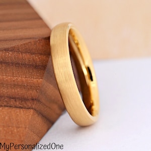 May include: A gold-colored ring with a brushed, matte finish and a smooth, rounded band. The ring is positioned at an angle, showcasing its metallic texture and circular shape. The text MyPersonalizedOne is printed in black at the bottom left.