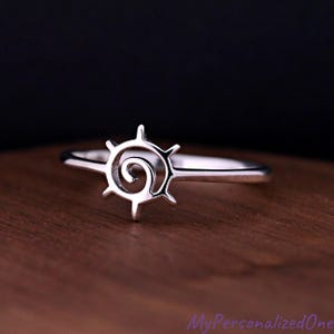 May include: A silver-toned ring featuring a central circular motif with a spiral design at its core and six radiating points extending outward. The band is thin and smooth, connecting to the sides of the circular emblem in a continuous metal loop.