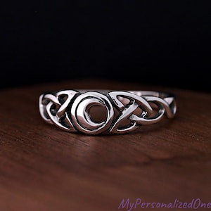 May include: A silver-colored ring with a Celtic knot design. The ring features interwoven patterns and a central crescent moon shape. The ring is displayed on a wooden surface, with a dark background.