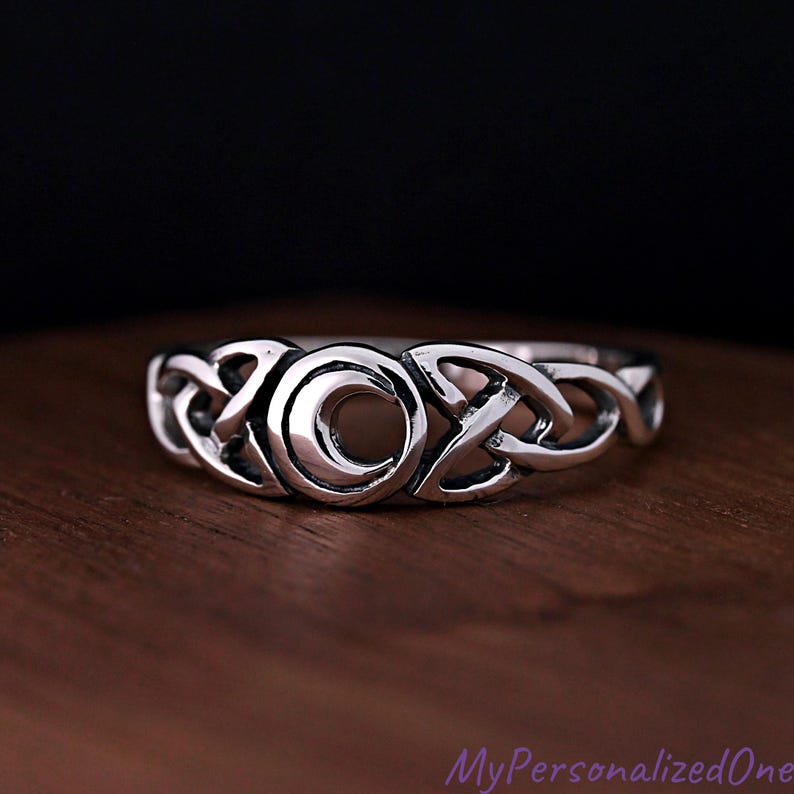 May include: A silver-toned metal ring featuring a central crescent moon motif flanked by intricate, continuous knotwork patterns. The band has a polished finish with darkened recessed areas that highlight the detailed design of the moon and knots.