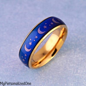 May include: A gold-toned band ring featuring a smooth, color-changing surface that transitions from blue to purple. The outer surface is decorated with a repeating pattern of light-colored crescent moons and stars. The text MyPersonalizedOne is visible.