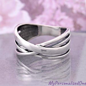 May include: A silver ring with a woven design. The ring features multiple bands that cross over each other, creating a textured and layered appearance. The metal has a polished finish, reflecting light and adding to its shine.