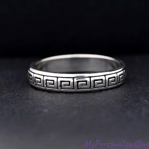 May include: A silver ring with a Greek key pattern around the band. The ring is displayed on a dark surface, showcasing the intricate design. The ring is a piece of jewelry.