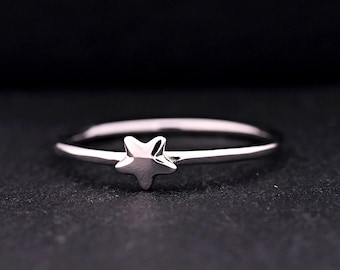 May include: A thin, polished silver-toned ring featuring a small, five-pointed star charm centered on the band. The metal surface reflects light, creating highlights on the star and the circular band against a dark, textured background.