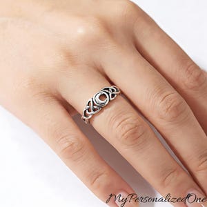 May include: A silver-toned ring featuring a central crescent moon motif flanked by intricate, interconnected triquetra knotwork patterns. The metal band has a polished finish with darkened recessed areas to highlight the detailed design elements.