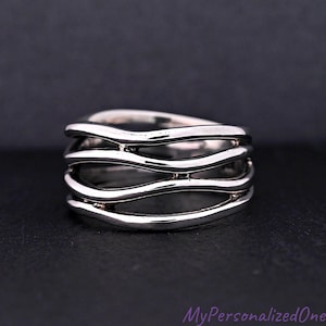 May include: A silver-colored ring with a wavy design. The ring features multiple bands that flow across the finger, creating a layered effect. The ring is set against a dark background.