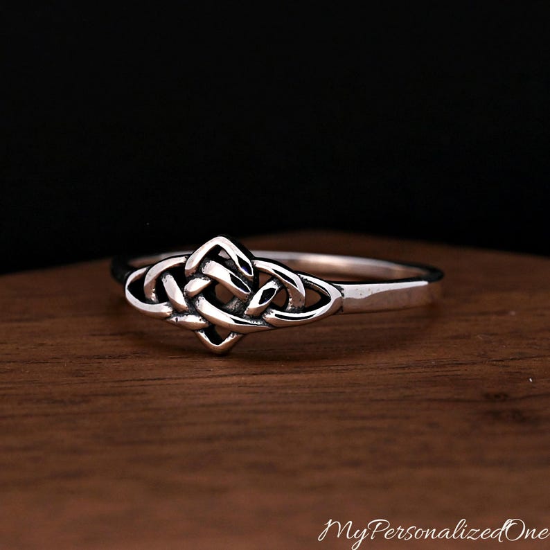 May include: A silver ring featuring a central Celtic knot design with intricate, polished metal loops. The band is smooth and tapers towards the back, showcasing a reflective finish that highlights the detailed, interwoven pattern of the knotwork.