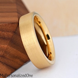 May include: A gold-toned ring with a brushed, matte exterior finish and a polished, reflective interior surface. The band features a flat profile with straight edges, showcasing a metallic texture that catches the light across its surface.