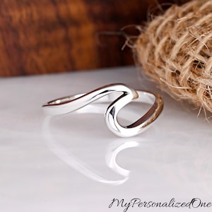 May include: A polished silver ring featuring a continuous, fluid wave-like design that curves upward to form a single crest. The band is smooth and reflective, showcasing a metallic sheen that catches the light, creating a clean and simple aesthetic.