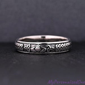 May include: A silver-colored ring with a Claddagh design, featuring a heart, crown, and hands, framed by Celtic knot patterns. The ring is set against a dark background, showcasing the intricate details of the jewelry.