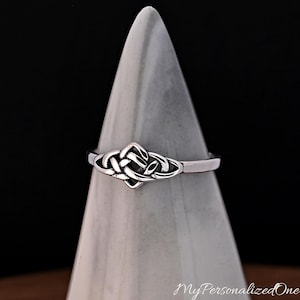 May include: A silver-coloured ring featuring a central knotwork design with intricate, interlaced loops. The metal has a polished finish with darkened recessed areas that highlight the woven pattern of the knot. The band is smooth and uniform.