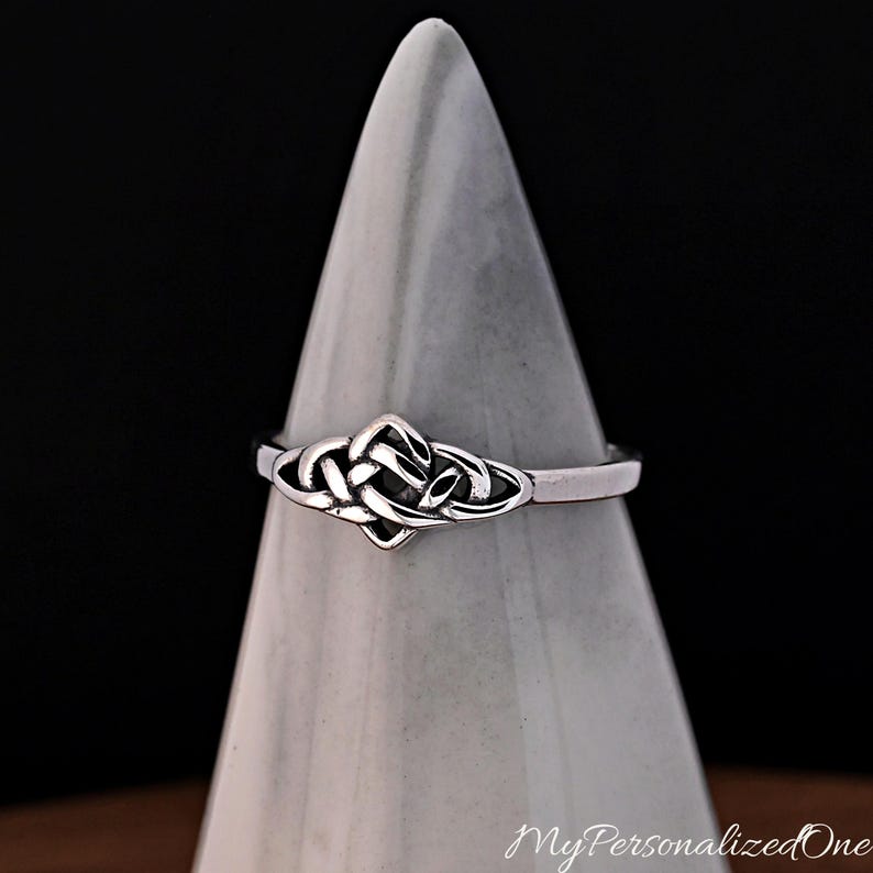 May include: A silver-coloured ring featuring a central knotwork design with intricate, interlaced loops. The metal has a polished finish with darkened recessed areas that highlight the woven pattern of the knot. The band is smooth and uniform.
