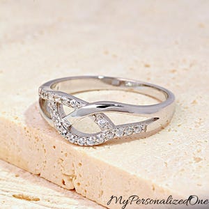 May include: A silver-colored ring with a unique, intertwined design. The ring features a band with a series of small, clear gemstones set along the top. The ring's design includes an open, interwoven pattern, creating an elegant and modern look. The ring is displayed on a light-colored surface.