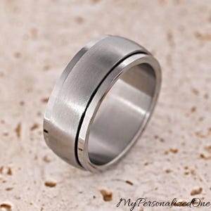 May include: A close-up of a silver-colored ring with a brushed metal finish. The ring has a wide band with a central rotating section. The ring is on a light-colored, textured surface. The text "MyPersonalizedOne" is visible.