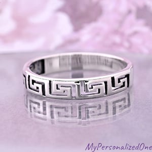 May include: A silver ring with a Greek key design. The ring is circular with a repeating geometric pattern around the band. The ring is displayed on a reflective surface with a soft, blurred background.