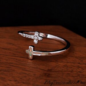 May include: A silver-colored ring with two cross designs. One cross is adorned with small, clear stones, while the other has an opal-like appearance. The ring is open-ended, allowing for size adjustment. The ring is on a wooden surface.