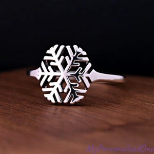 May include: A silver snowflake ring. The ring features a detailed, openwork design of a snowflake. The snowflake is the focal point, with intricate patterns and cutouts. The ring is set against a wooden surface with a dark background.
