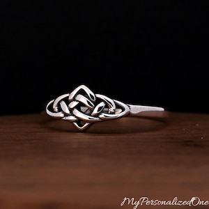 May include: A silver ring featuring a central knotwork design with interlacing bands. The metal has a polished finish with darkened recessed areas that highlight the intricate woven pattern of the knot. The band is smooth and tapers towards the back.