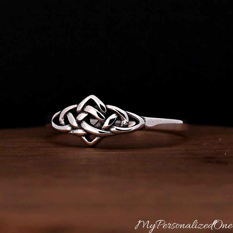 May include: A silver ring featuring a central knotwork design with interlacing bands. The metal has a polished finish with darkened recessed areas that highlight the intricate woven pattern of the knot. The band is smooth and tapers towards the back.