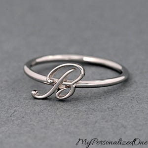 May include: A silver-toned metal ring featuring a cursive letter B as the central focal point. The band is thin and smooth, connecting to the stylized script initial. The design is clean and polished, showcasing a metallic finish throughout the piece.
