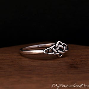 May include: A silver ring featuring a polished band and a central knot design with oxidised detailing in the recessed areas. The metal has a smooth, reflective finish, and the intricate knotwork creates a symmetrical pattern on the top of the ring.