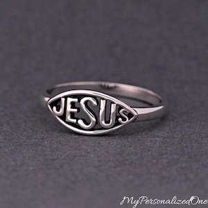May include: A silver ring with the word "JESUS" cut out in a stylized font. The ring has a smooth, polished finish and is set against a dark gray background. The ring's design is simple yet elegant.