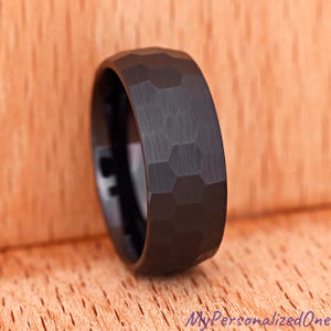 May include: A black tungsten ring with a hammered, hexagonal pattern. The ring has a polished interior and a textured exterior. The ring is a dark charcoal color and is set against a wooden background.