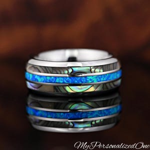 May include: A silver-toned band ring featuring a central inlay of bright blue crushed opal flanked by two strips of iridescent abalone shell. The ring has a polished metallic finish and the text MyPersonalizedOne is visible at the bottom right.