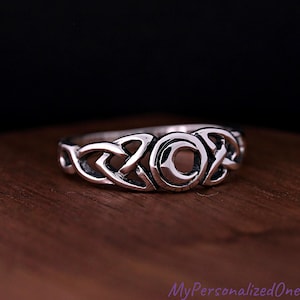 May include: A silver-toned metal ring featuring a central crescent moon motif flanked by intricate, continuous Celtic knotwork patterns. The band has a polished finish with darkened recessed areas that highlight the detailed metalwork design.