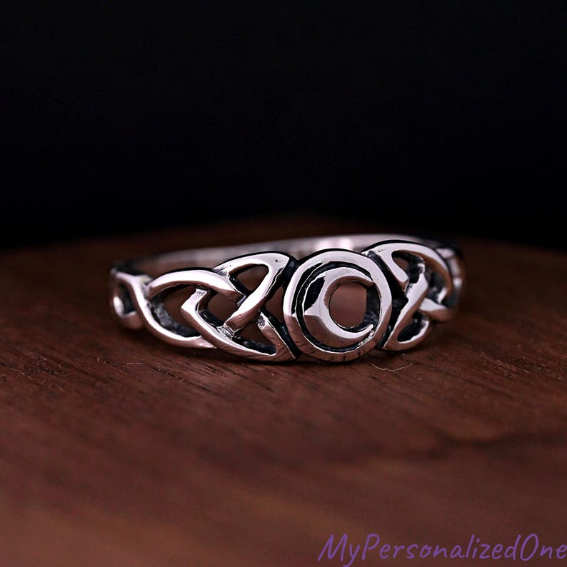 May include: A silver-toned metal ring featuring a central crescent moon motif flanked by intricate, continuous Celtic knotwork patterns. The band has a polished finish with darkened recessed areas that highlight the detailed metalwork design.