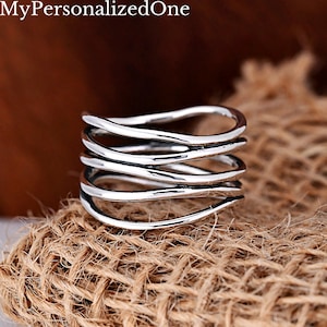 May include: A silver-toned ring featuring a multi-layered design with five distinct, wavy, interconnected bands that wrap around the finger. The metal has a polished finish with darkened recessed areas to highlight the fluid, organic curves of the structure.