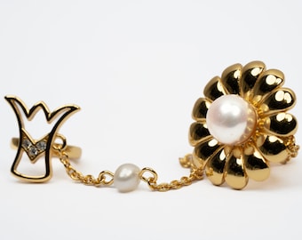 May include: A gold ring set with a pearl and a flower design, connected to a second ring with a crown design by a gold chain with a pearl charm.
