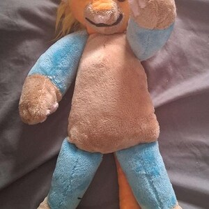May include: A plush toy featuring an orange head with yellow hair, blue ears, and black eyes. The body is tan with blue arms and legs, and tan paws. A long orange tail with a yellow tuft of hair extends from the bottom of the plush creature.