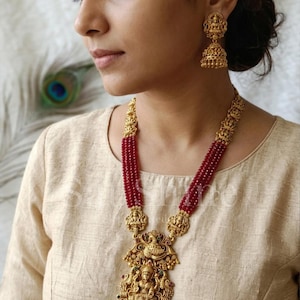 May include: A gold-coloured necklace and matching earrings set. The necklace features a large pendant with intricate carvings, red beaded strands, and a detailed deity figure. The earrings have a similar design. The jewellery is worn by a person wearing a beige top.