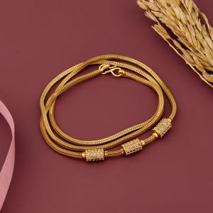 Peut inclure: Un collier doré avec un design de chaîne en maille. Le collier comporte trois accents cylindriques sertis de diamants. Le collier est posé sur une surface marron avec une brindille de blé et un ruban rose en arrière-plan.