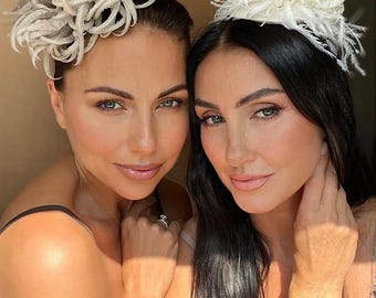 May include: Two women wearing decorative headpieces. One headpiece is a large, textured, light gray design. The other is a smaller, white, feathery design. Both women have fair skin and are smiling.