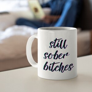 May include: White ceramic coffee mug with the text "still sober bitches" in black with a glitched effect.