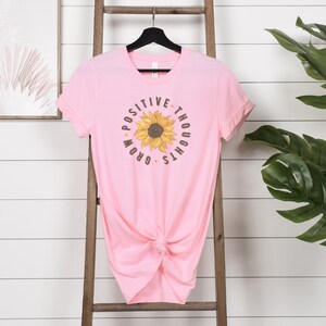 May include: Pink t-shirt with a sunflower graphic and the text "Positive Thoughts Grow".