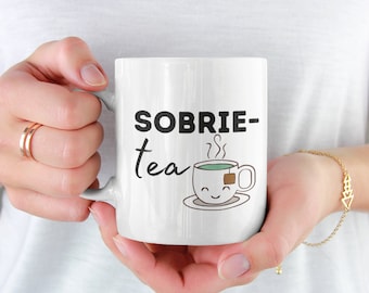 May include: White ceramic mug with black text that reads "SOBRIE-tea" and a cartoon illustration of a teacup with a tea bag and steam coming out of it.